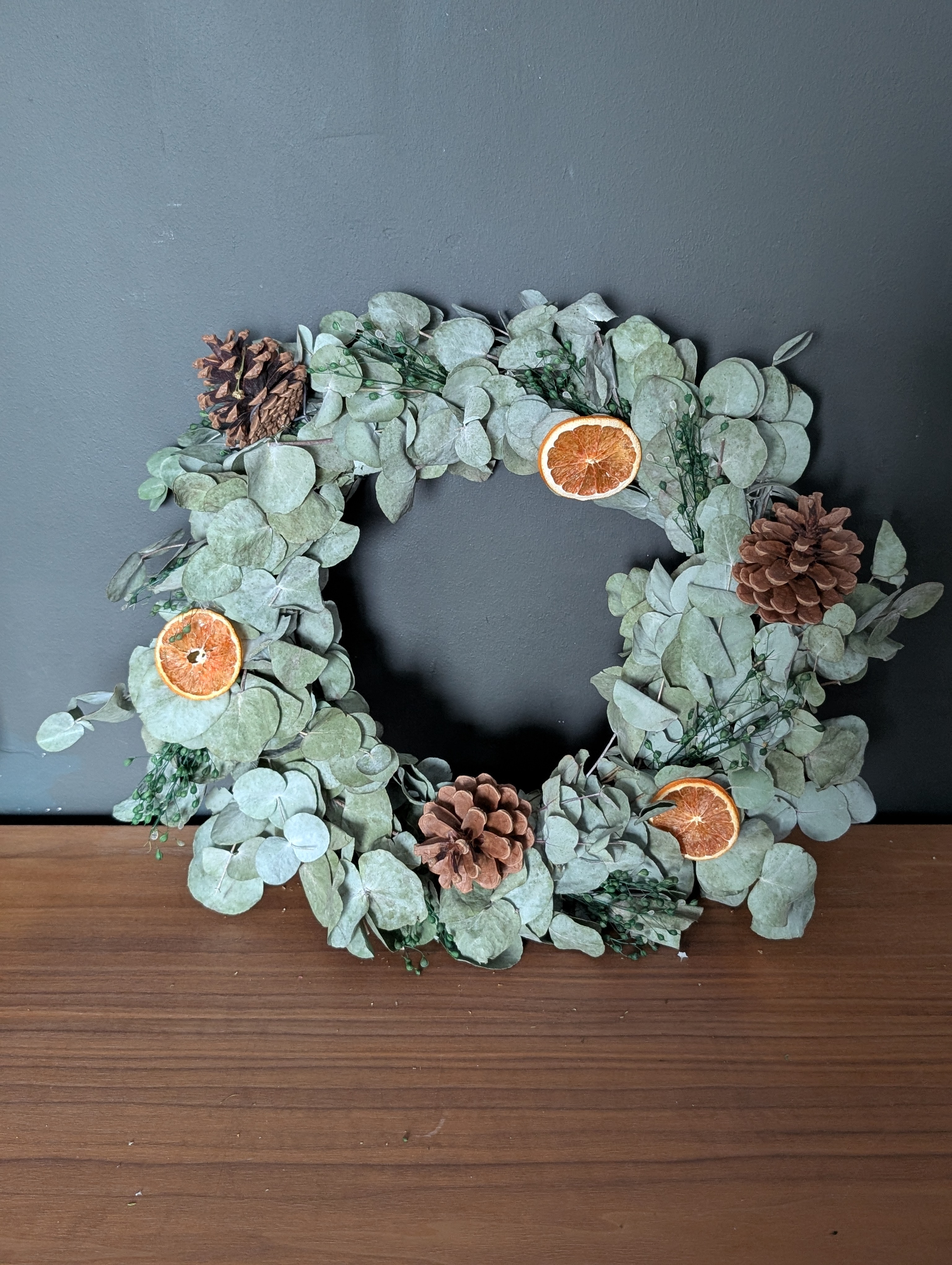 A circular wreath made of dried eucalyptus leaves, accented with pinecones and slices of dried orange. The wreath is displayed against a dark grey wall on a wooden surface, creating a rustic and natural look.