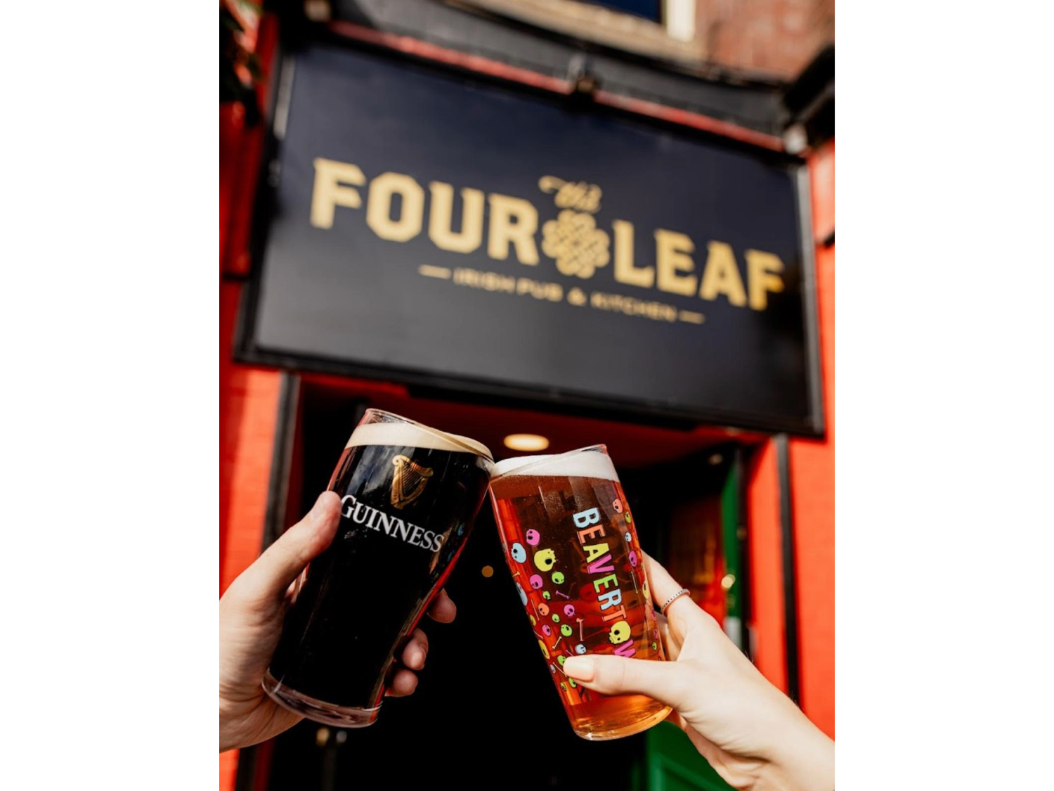 A pint of beer and a pint of Guinness being clinked together under The Four Leaf Irish Bar & Kitchen's sign. 