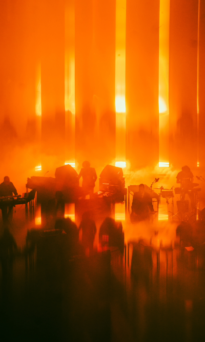 Silhouetted musicians perform on stage surrounded by dense orange fog and dramatic vertical light beams. The scene is bathed in warm, glowing tones, creating an abstract, atmospheric effect with blurred reflections on the floor.