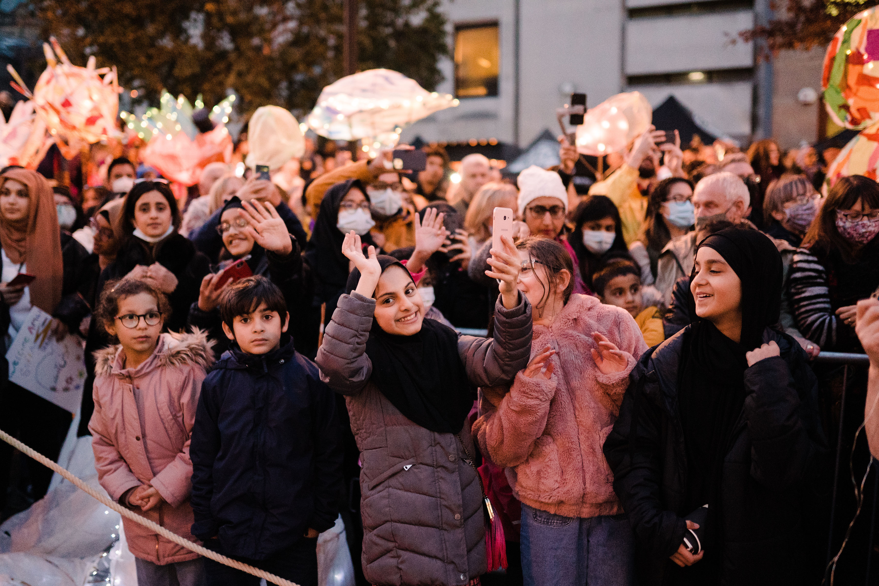 Crowd gathered at an outdoor event during dusk, with people standing behind a rope barrier and raising hands or holding phones. Illuminated lanterns and lighted decorations are visible in the background, along with buildings and trees.