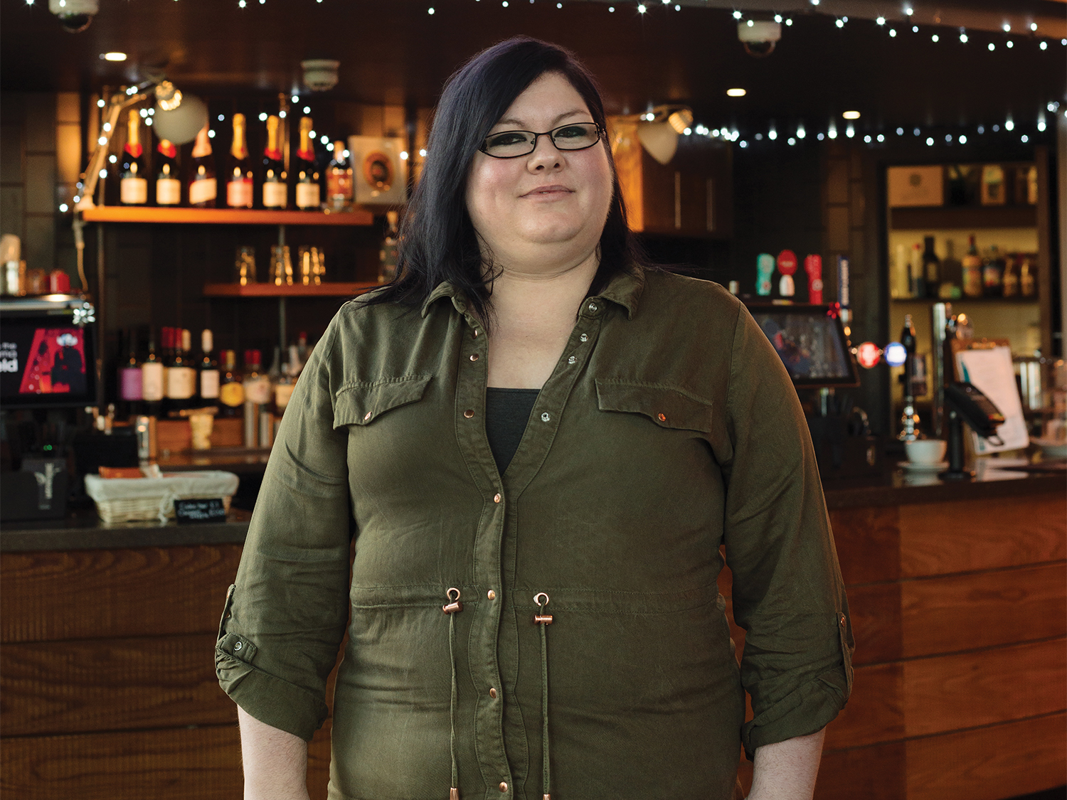 A person wearing an olive green button-up shirt is standing indoors in front of a wooden bar counter. The background features shelves stocked with bottles, a coffee machine, and string lights hanging across the top, creating a warm and inviting atmosphere.