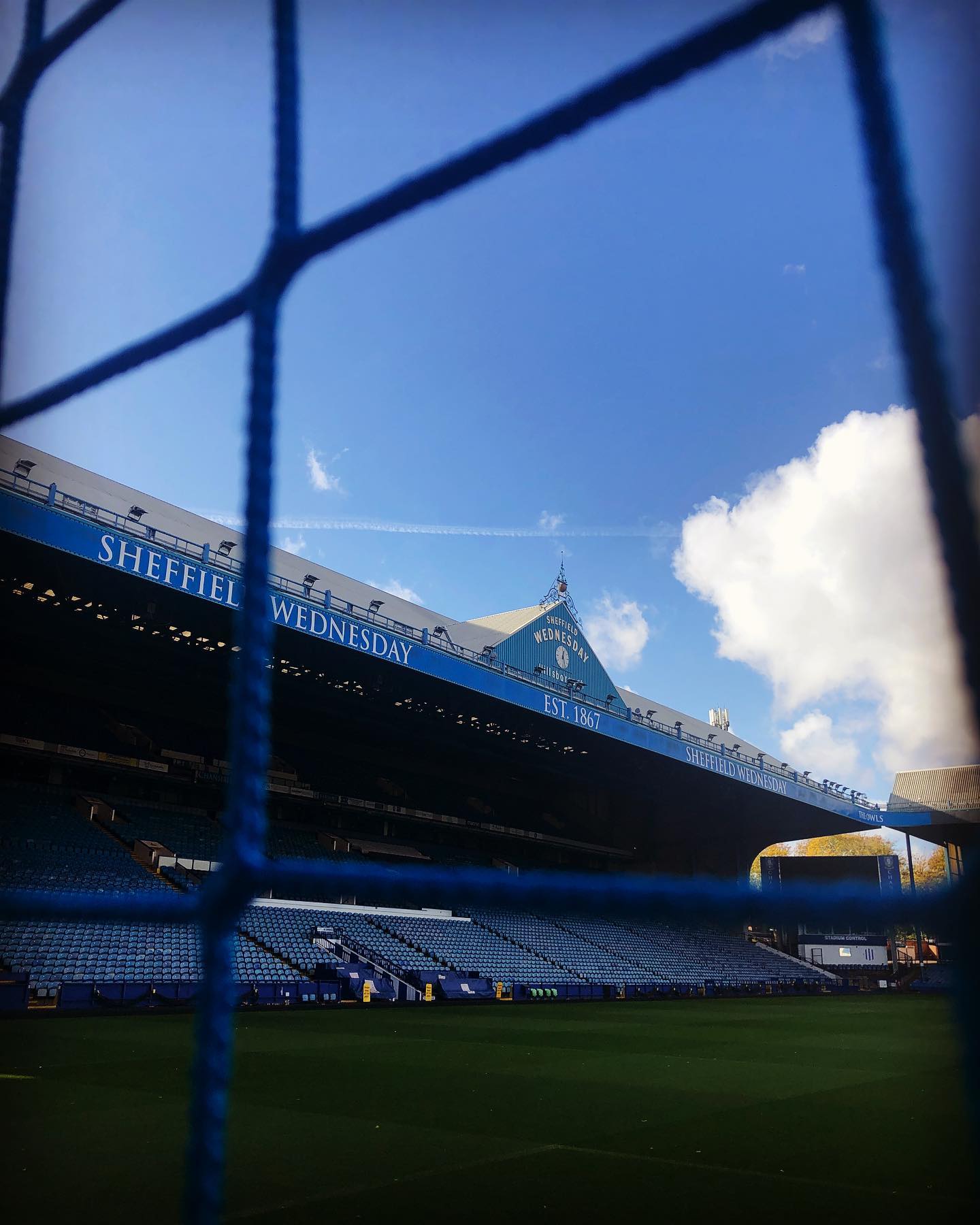 View of Hillsborough Stadium, home of Sheffield Wednesday FC, taken from behind a goal net. The blue seating and main stand are visible, with signage reading ‘Sheffield Wednesday’ and ‘Est. 1867’ under a bright blue sky with scattered clouds.