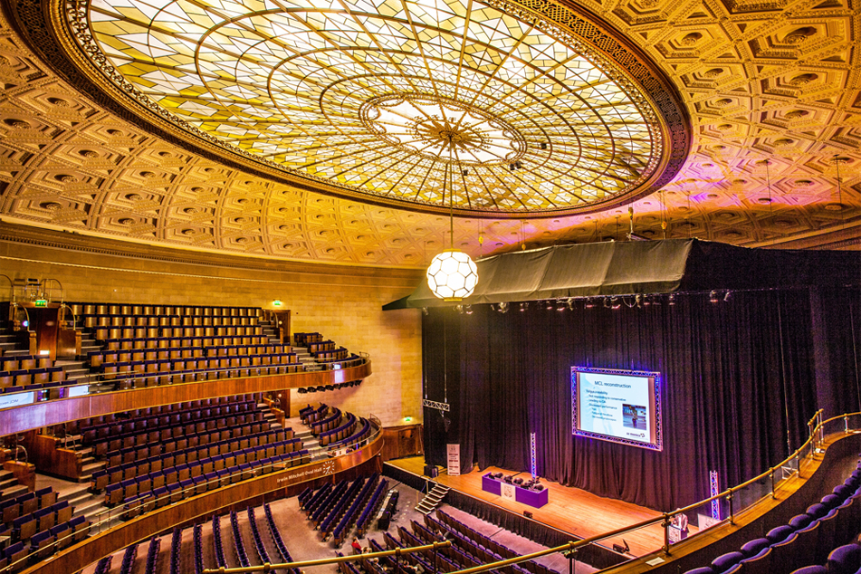 The Oval Hall at Sheffield City Hall. Grand auditorium with tiered seating and a large stage featuring a projection screen. The ornate ceiling has an intricate geometric design with a central stained-glass dome and a hanging light fixture. The stage is framed by black curtains, and the seating area includes multiple balconies with curved railings.