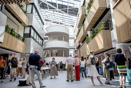 Spacious modern atrium with a glass ceiling and natural light, featuring multiple floors with wooden slatted balconies and green plants. A group of people is gathered in the open central area, some standing near tables with refreshments. The design includes curved white staircases and sleek architectural details, creating a bright and contemporary setting.