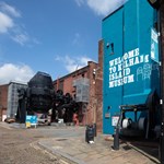 The entrance to the Kelham Island Museum. On the right is a blue-painted wall with 'Welcome To Kelham Island Museum' on it. In the background you can see a huge Bessemer Converter that was used in the production of steel.