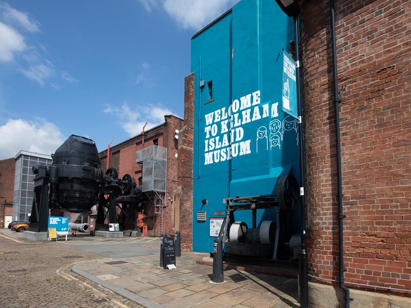 The entrance to the Kelham Island Museum. On the right is a blue-painted wall with 'Welcome To Kelham Island Museum' on it. In the background you can see a huge Bessemer Converter that was used in the production of steel.