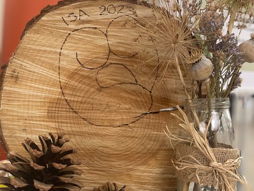 A decorative display featuring a large wooden tree slice with visible growth rings and an engraved design labeled “ISA 2023.” The slice is surrounded by natural elements, including dried flowers, seed heads, pinecones, and a glass jar wrapped with burlap and twine, creating a rustic and organic arrangement.