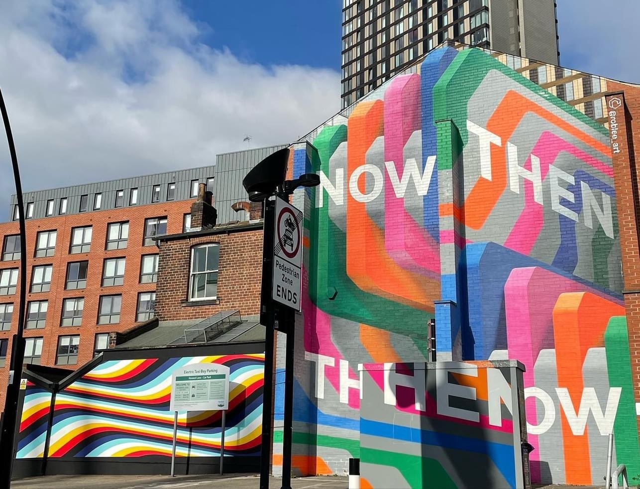 Street art murals on adjacent buildings in an urban setting. One mural features bold, colorful geometric shapes with large white text reading ‘NOW THEN.’ The other mural displays flowing rainbow-colored stripes across a wall. Modern buildings and a tall tower block are visible in the background under a partly cloudy sky.