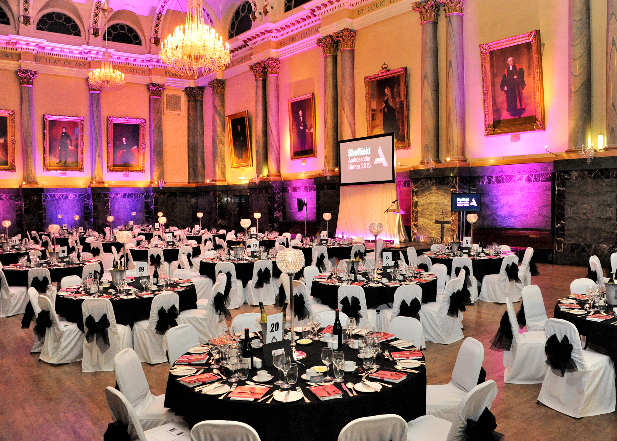 Elegant banquet hall with high ornate ceilings, chandeliers, and large portraits on the walls. Round tables are set for a formal dinner with white chair covers tied with black bows, black tablecloths, and red menus. A projector screen at the front displays “Sheffield Ambassador Dinner 2015.” Warm lighting with pink and orange uplights creates a vibrant atmosphere.