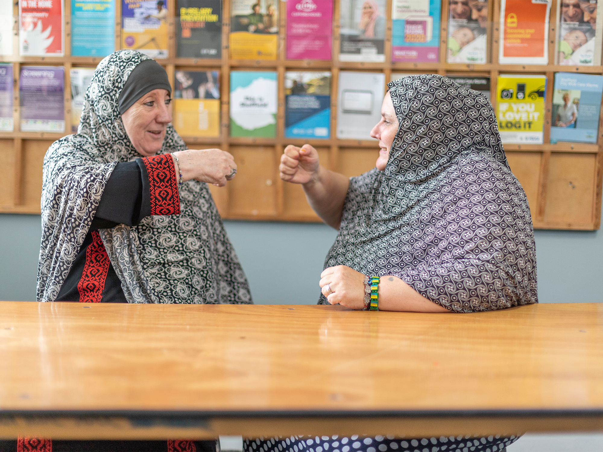 Two people sitting at a wooden table, engaged in conversation and gesturing with their hands. Both are wearing patterned shawls. Behind them is a wall-mounted display board filled with colourful posters and flyers arranged in a grid.