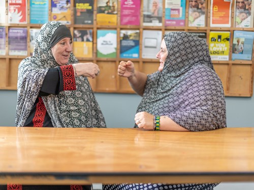 Two people sitting at a wooden table, engaged in conversation and gesturing with their hands. Both are wearing patterned shawls. Behind them is a wall-mounted display board filled with colourful posters and flyers arranged in a grid.