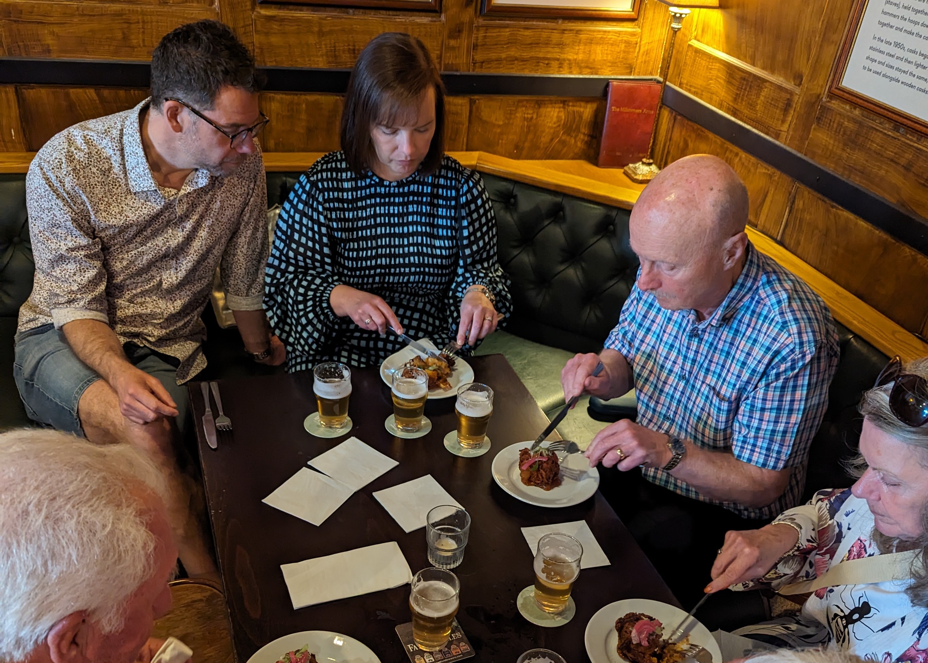 Group of people sitting around a wooden table in a pub, eating plated meals and drinking beer. The table has several glasses of beer, plates of food, and napkins, with wooden panelling and cushioned seating in the background.