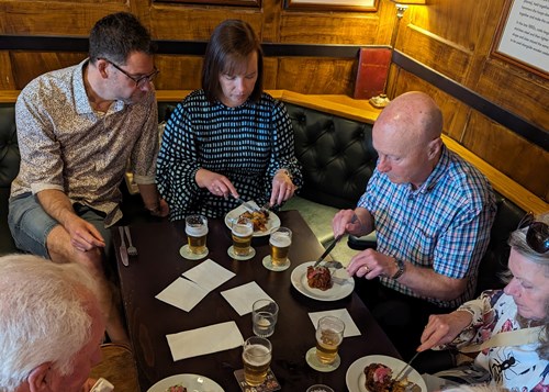 Group of people sitting around a wooden table in a pub, eating plated meals and drinking beer. The table has several glasses of beer, plates of food, and napkins, with wooden panelling and cushioned seating in the background.