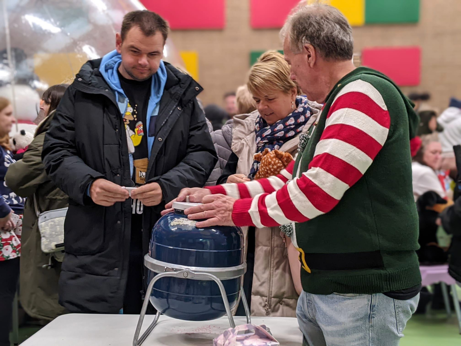 Indoor festive event with a group of people gathered around a blue raffle drum or tombola on a table. One person is wearing a green and red striped Christmas sweater and appears to be operating the drum, while others stand nearby, possibly buying tickets. The background shows colourful wall panels and more attendees in a busy hall decorated for the holidays.