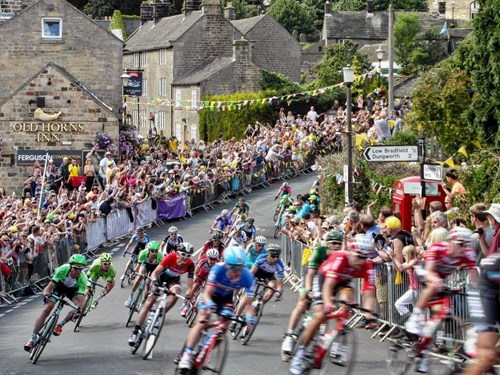 Cyclists racing through a sharp bend in a village street lined with stone buildings, including the Old Horns Inn. Large crowds of spectators stand behind metal barriers, and colourful bunting decorates the route.