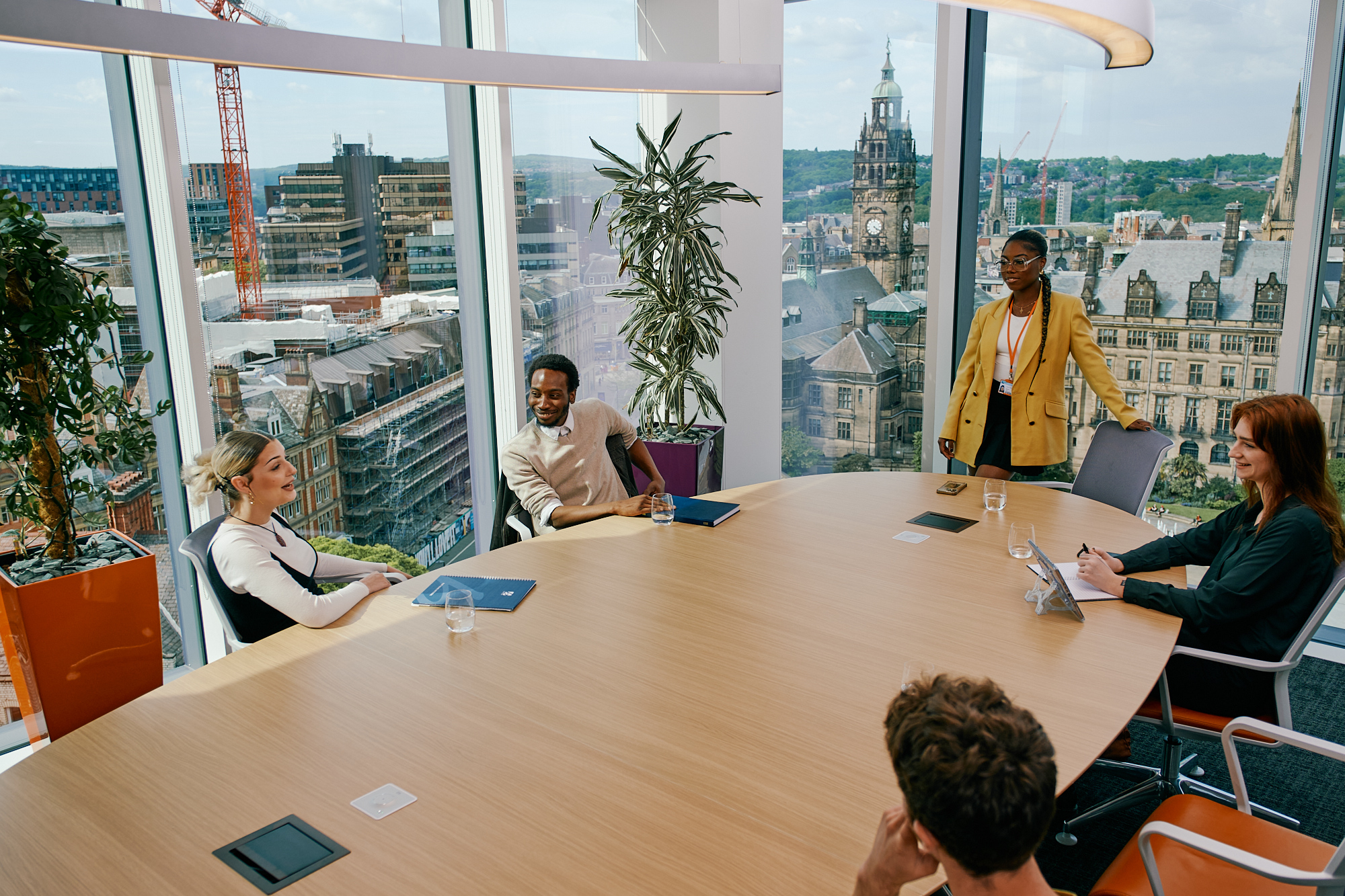 Four people, sat around a table in an office, are having a meeting. Through the pane glass windows Sheffield city centre is visible. 