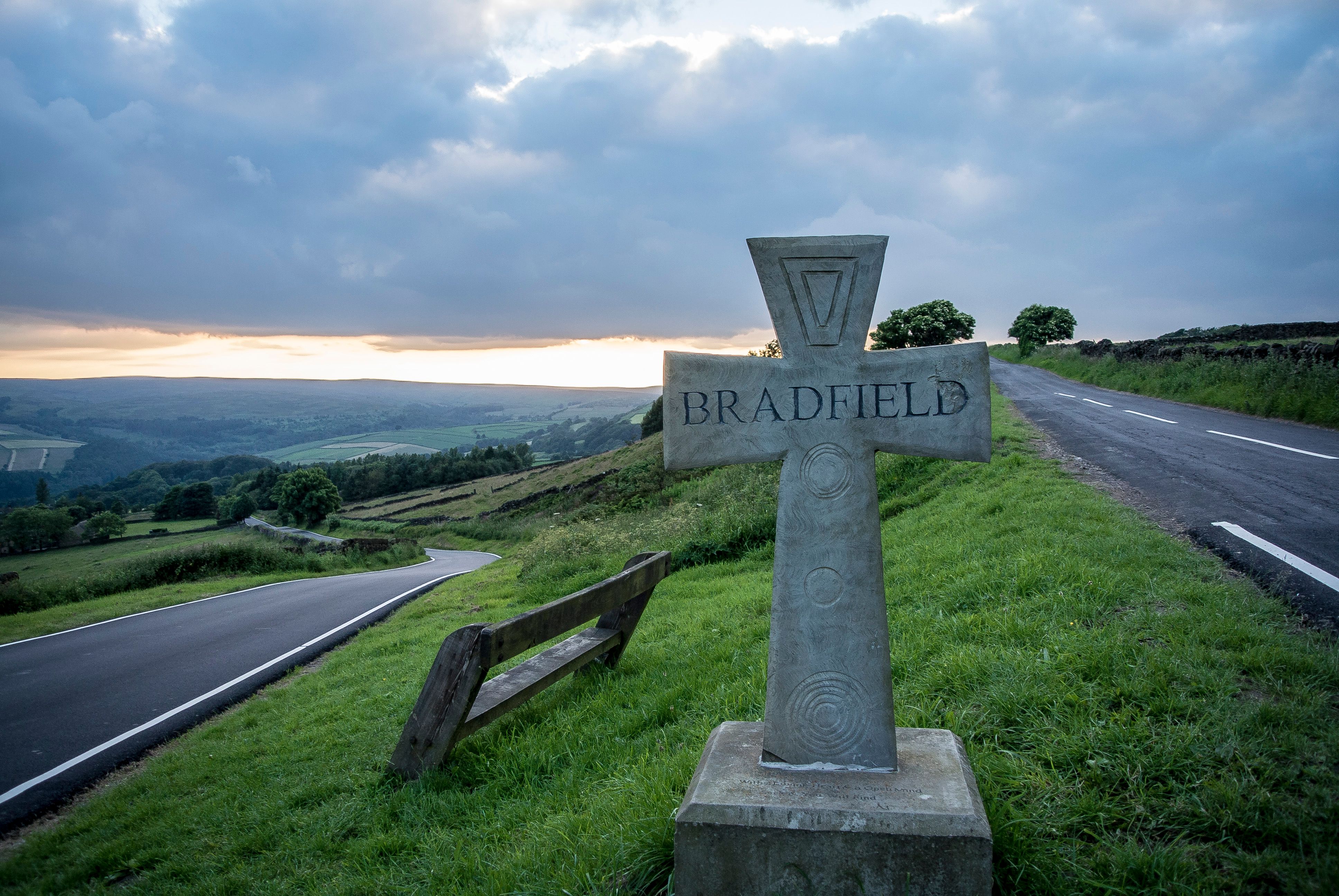 On a grass verge between two roads stands a stone cross with the word 'Bradfield' carved into it.