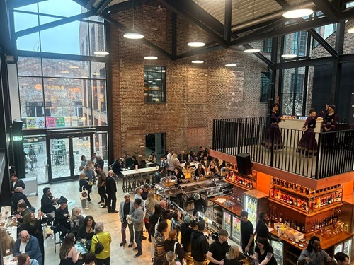 Interior of a large industrial-style bar and restaurant with exposed brick walls, high ceilings, and large windows. The space is filled with people dining and socializing at tables and around a central bar stocked with bottles and glassware. A mezzanine level overlooks the main floor, where three performers in matching outfits stand near the railing. Bright lighting and modern decor create a lively atmosphere.