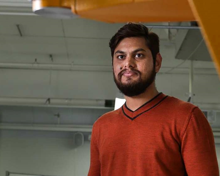 A person wearing an orange long-sleeve sweater with a black V-neck trim stands indoors near large industrial equipment. The background shows a high ceiling with exposed pipes and light fixtures, suggesting a factory or workshop setting.