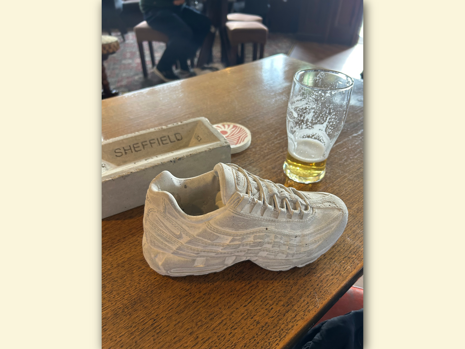 A concrete sneaker sculpture placed on a wooden table next to a half-empty pint glass. Behind the shoe is a rectangular concrete block with the word “SHEFFIELD” engraved on it. The setting appears to be indoors, possibly a pub, with chairs and patterned carpet visible in the background.
