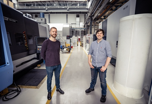 Two people standing in a spacious industrial workshop with large machinery on both sides. The environment features high ceilings, overhead beams, and bright lighting. The floor has marked yellow lines for pathways, and various equipment and workstations are visible in the background.