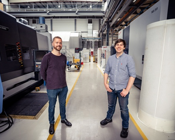 Two people standing in a spacious industrial workshop with large machinery on both sides. The environment features high ceilings, overhead beams, and bright lighting. The floor has marked yellow lines for pathways, and various equipment and workstations are visible in the background.