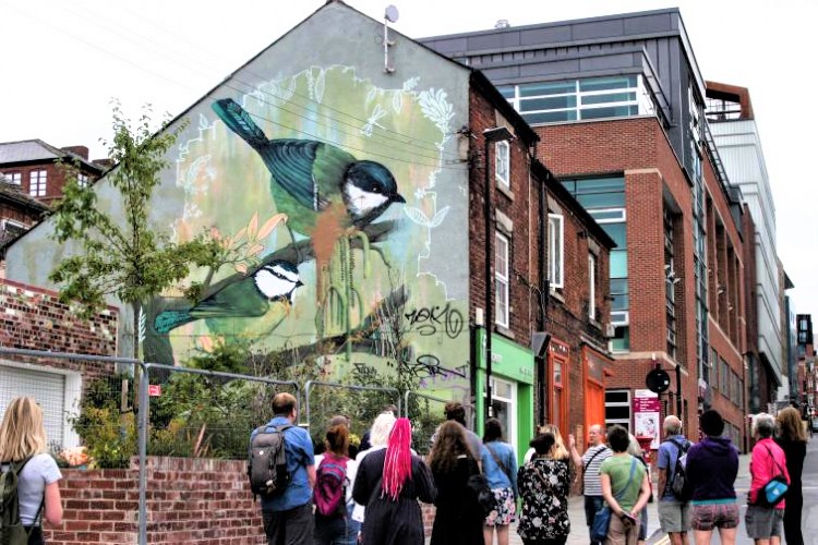 Group of people walking down an urban street toward a building with a large mural of two birds perched on branches, painted on the side wall. The mural features natural tones of green and brown, and the surrounding area includes brick buildings, a green shopfront, and modern architecture in the background.