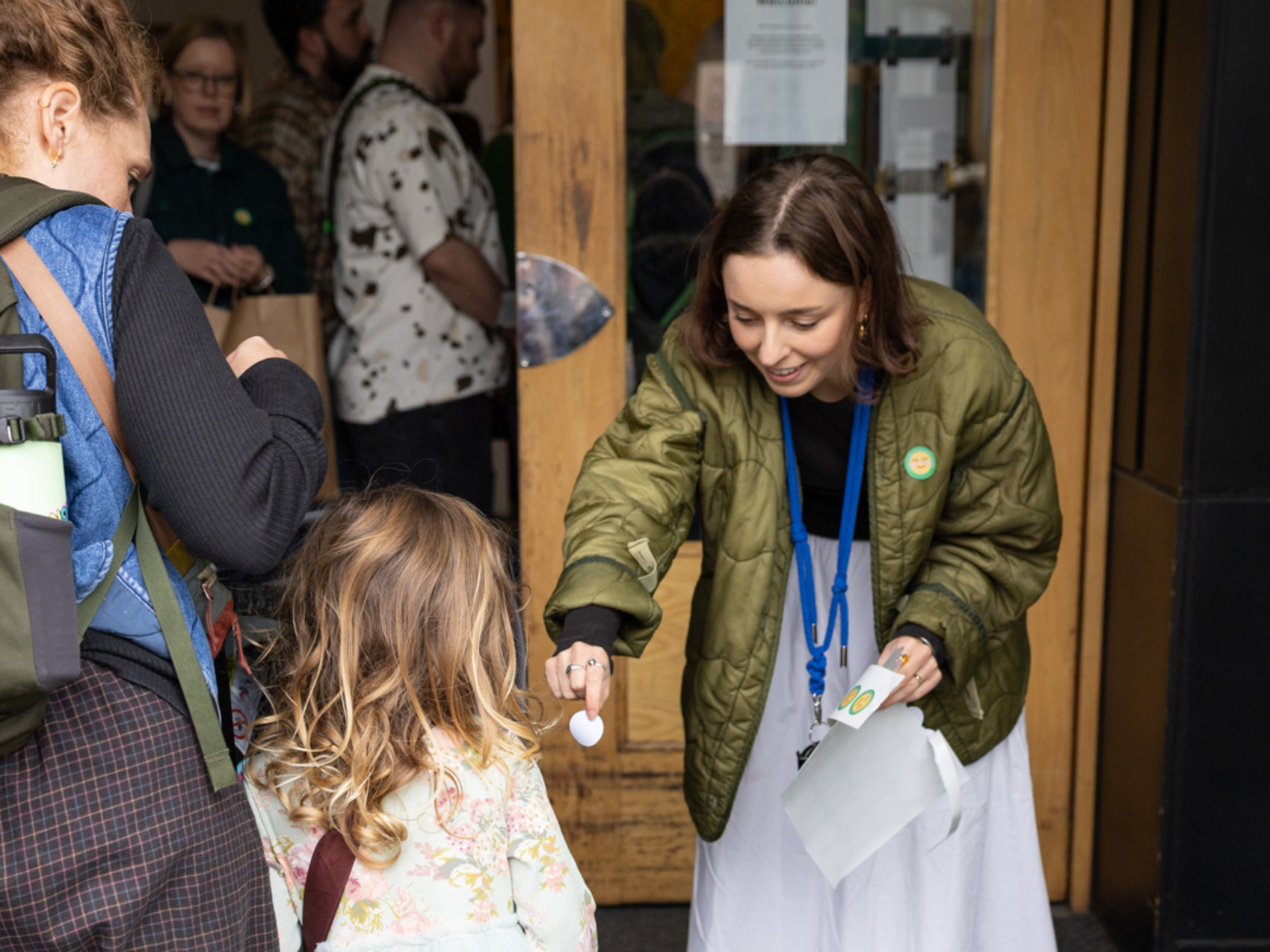 A person in a green jacket bends down to hand an object to a child with long, wavy hair, who is accompanied by another person wearing a backpack. Several people stand nearby at the entrance of a building, observing the interaction.