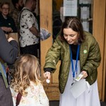 A person in a green jacket bends down to hand an object to a child with long, wavy hair, who is accompanied by another person wearing a backpack. Several people stand nearby at the entrance of a building, observing the interaction.