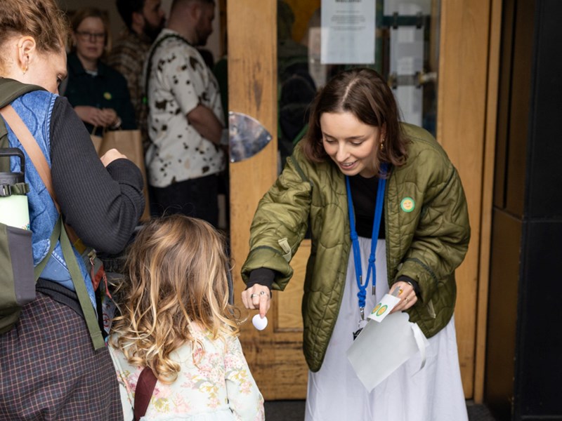 A person in a green jacket bends down to hand an object to a child with long, wavy hair, who is accompanied by another person wearing a backpack. Several people stand nearby at the entrance of a building, observing the interaction.