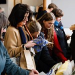 A group of people dressed in warm clothing browse and examine textiles or fabric items at a market or store. The scene captures a moment of focused shopping activity, with individuals closely inspecting the products on display.