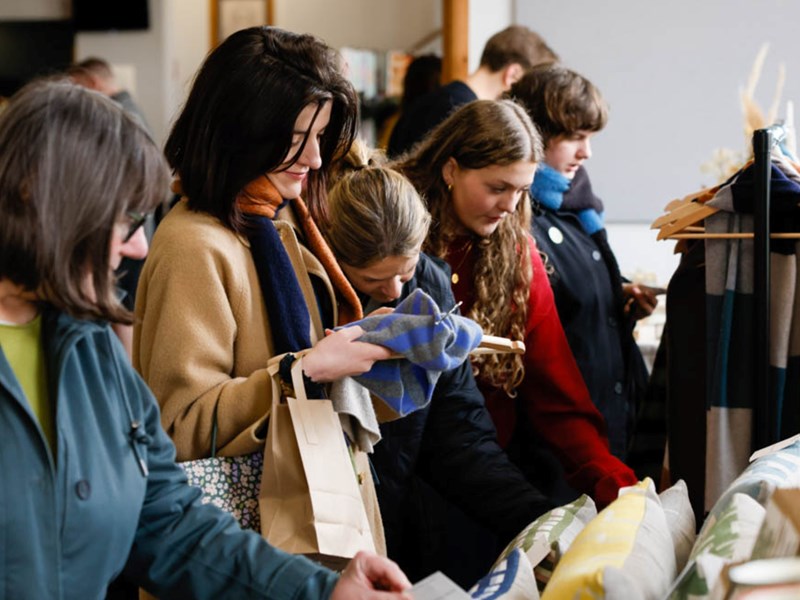 A group of people dressed in warm clothing browse and examine textiles or fabric items at a market or store. The scene captures a moment of focused shopping activity, with individuals closely inspecting the products on display.