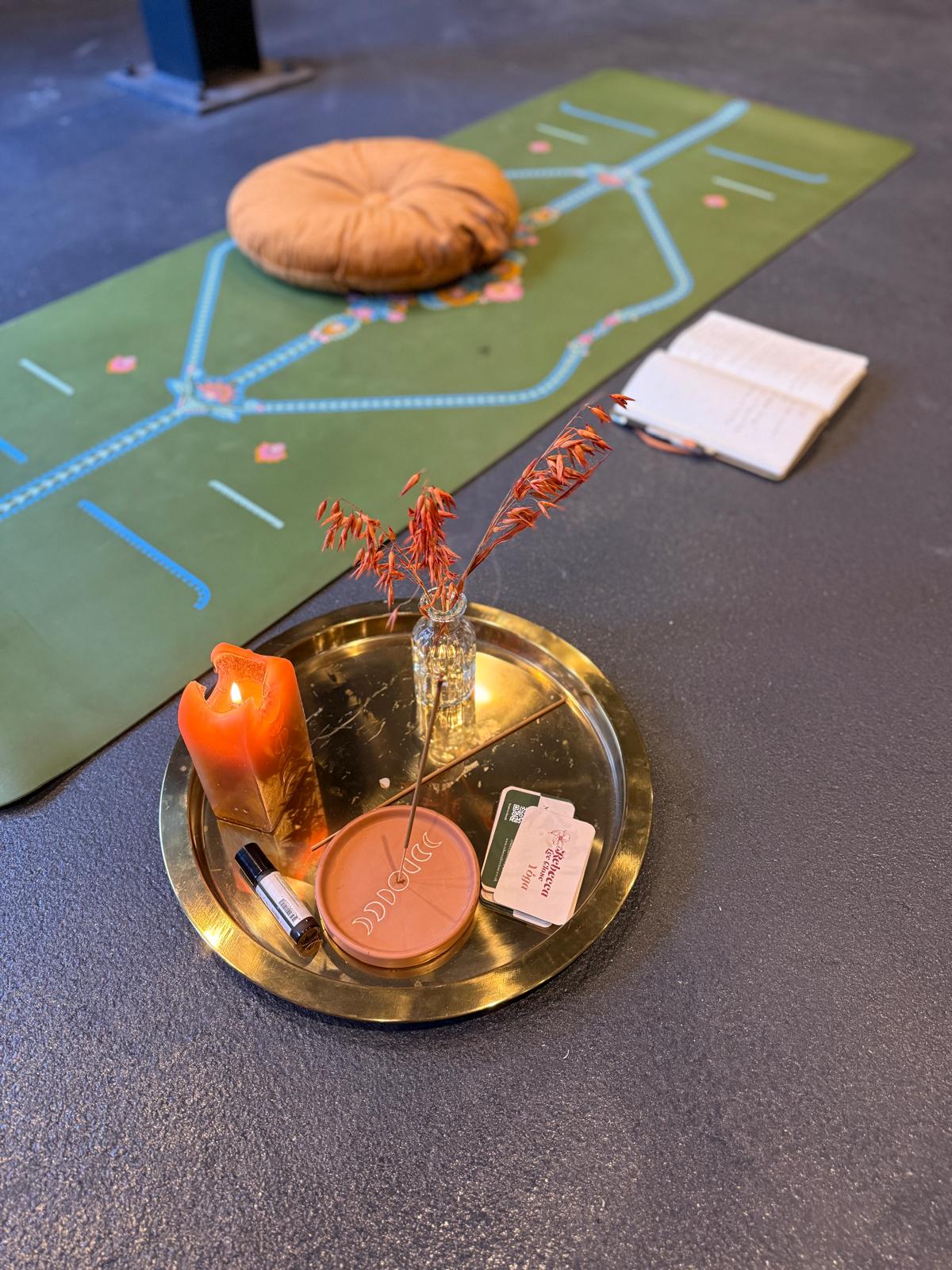 A brass tray on a dark floor holds an orange candle, a small glass vase with dried red stems, an incense stick in a round holder, a lip balm tube, and two cards. Behind the tray is a green mat with blue and pink patterns, a round tan cushion, and an open notebook with writing.
