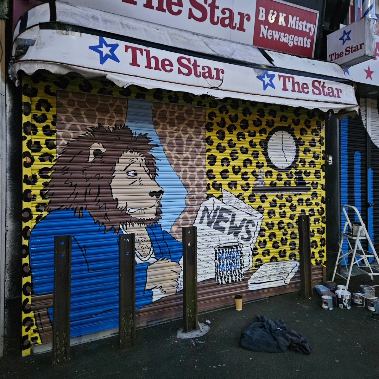 A shopfront with a closed shutter painted in a bold mural. The artwork shows a cartoon-style lion wearing a blue jacket, sitting at a table with a newspaper labeled “NEWS” and a mug. The background features a yellow leopard-print pattern and a brown curtain-like design, with a round white clock on the wall. Paint cans and a ladder are visible on the ground, suggesting the mural is being completed.
