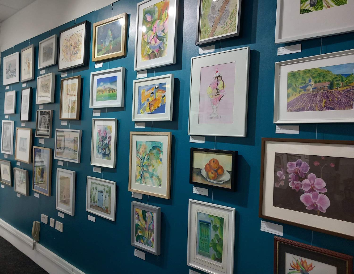 A gallery wall displaying a variety of framed artworks in different styles and sizes. The pieces include colorful paintings of flowers, landscapes, abstract designs, and still life, all arranged neatly on a teal-coloured wall. Small labels are visible beneath each frame, indicating titles or artist information.