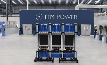 ITM Power: Industrial interior with a large blue and gray modular unit positioned in the center of a spacious factory floor. Behind it is a dark blue wall featuring the ITM Power logo in white. The space has high ceilings with metal beams, bright overhead lighting, and multiple doors and potted plants along the walls.