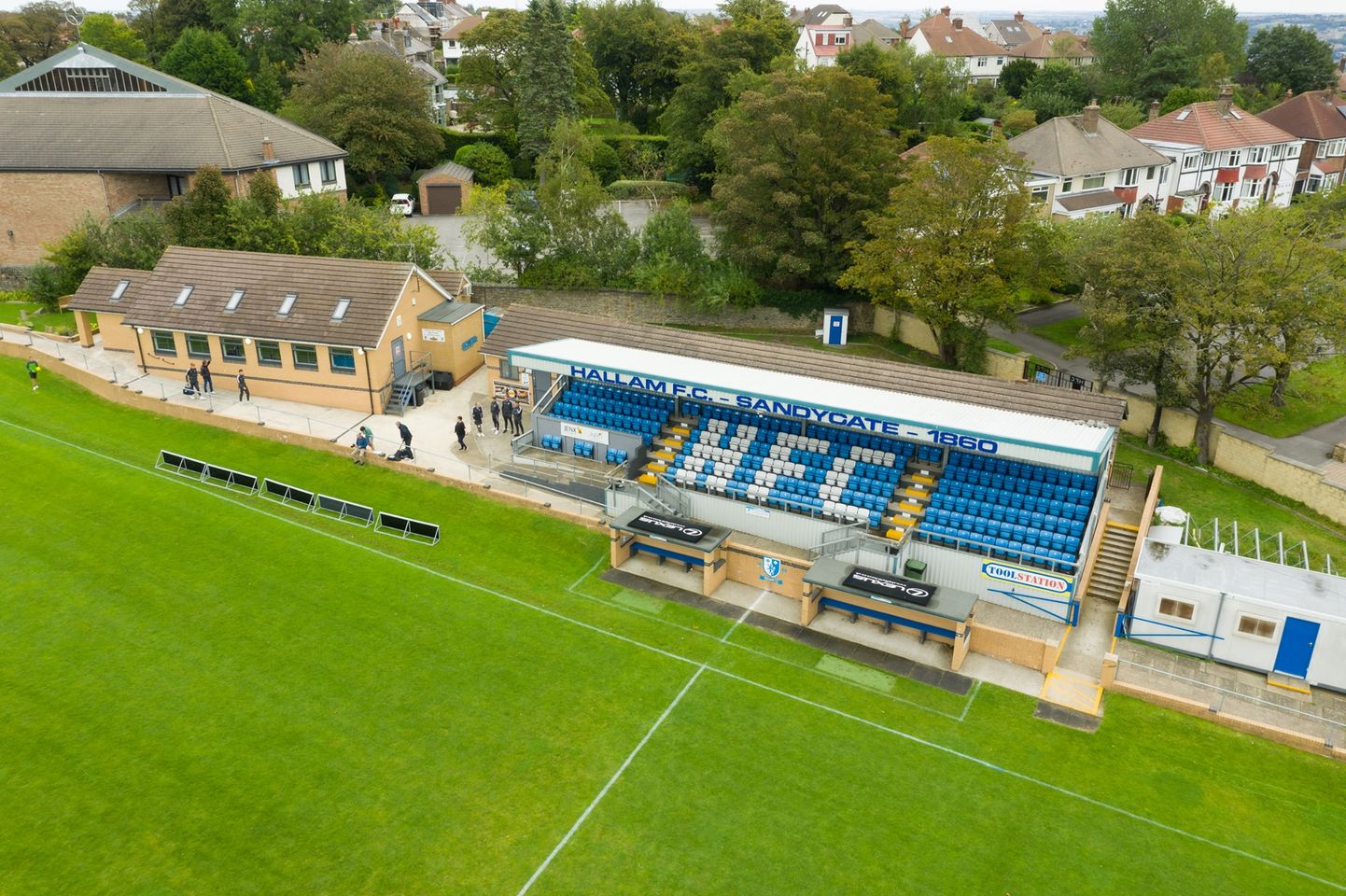 Aerial view of Hallam FC’s Sandygate stadium showing the main stand with blue seating and signage reading ‘Hallam F.C. Sandygate 1860.’ Below the stand are two dugouts, and to the left is a clubhouse building. The pitch is in the foreground, with trees and residential houses in the background.