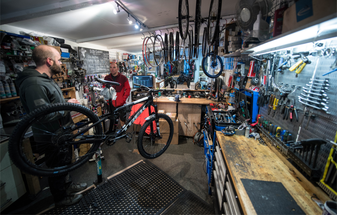 A photograph of the interior of The Bike Garage with the walls covered in tools and spare parts. There are two men working on bikes.