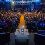 A large audience is sat waiting for an event to begin. On the stage are two armchairs on either side of a low table that has two glasses and a pitcher of water sat on it.