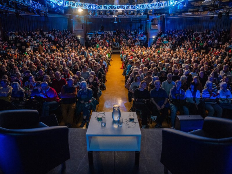 A large audience is sat waiting for an event to begin. On the stage are two armchairs on either side of a low table that has two glasses and a pitcher of water sat on it.