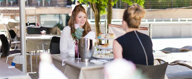 Two people sit at an outdoor café table, conversing. The table includes a flower vase, a metal ice bucket, and plates of food. Other patrons are visible in the background among tables and chairs, with trees providing shade in a relaxed, accessible setting.