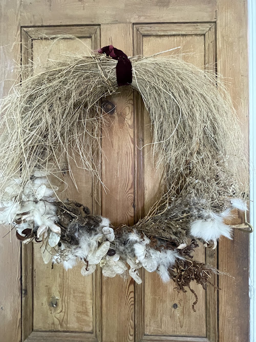 A natural wreath crafted from dried grasses, seed pods, and fluffy white plant fibers, arranged in a loose, organic style. It hangs on a wooden door and is finished with a simple dark velvet ribbon at the top.