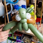 A person holds a balloon sculpture of a turtle made from green, white, and brown balloons, featuring large eyes and a small smile. In the background, other balloon sculptures and decorations are visible, suggesting an indoor event focused on balloon art.