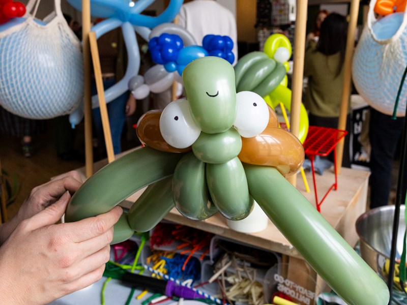 A person holds a balloon sculpture of a turtle made from green, white, and brown balloons, featuring large eyes and a small smile. In the background, other balloon sculptures and decorations are visible, suggesting an indoor event focused on balloon art.