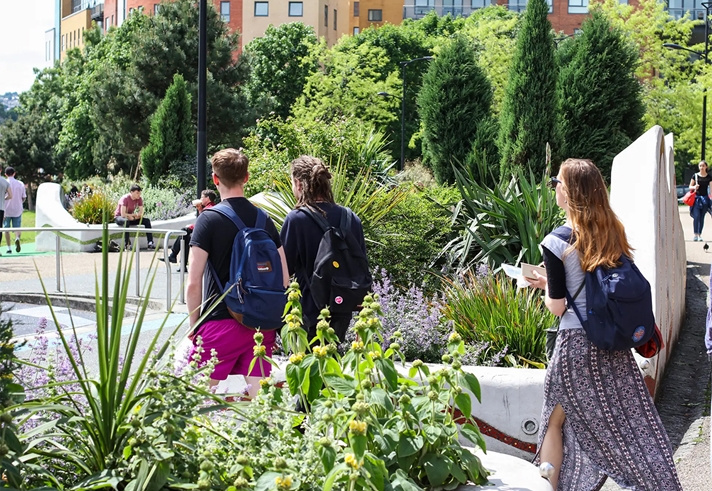 People walking through a public space, in an urban setting, that is full of trees and plants.