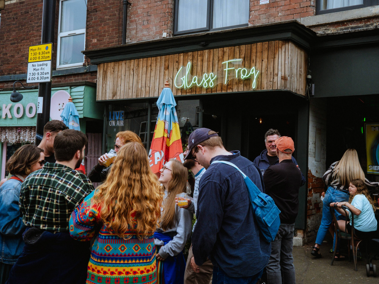Group of people gathered outside a venue with a wooden facade and a neon sign reading ‘Glass Frog’. The entrance is flanked by colorful umbrellas, and the setting appears to be a lively street with adjacent shops and signage.