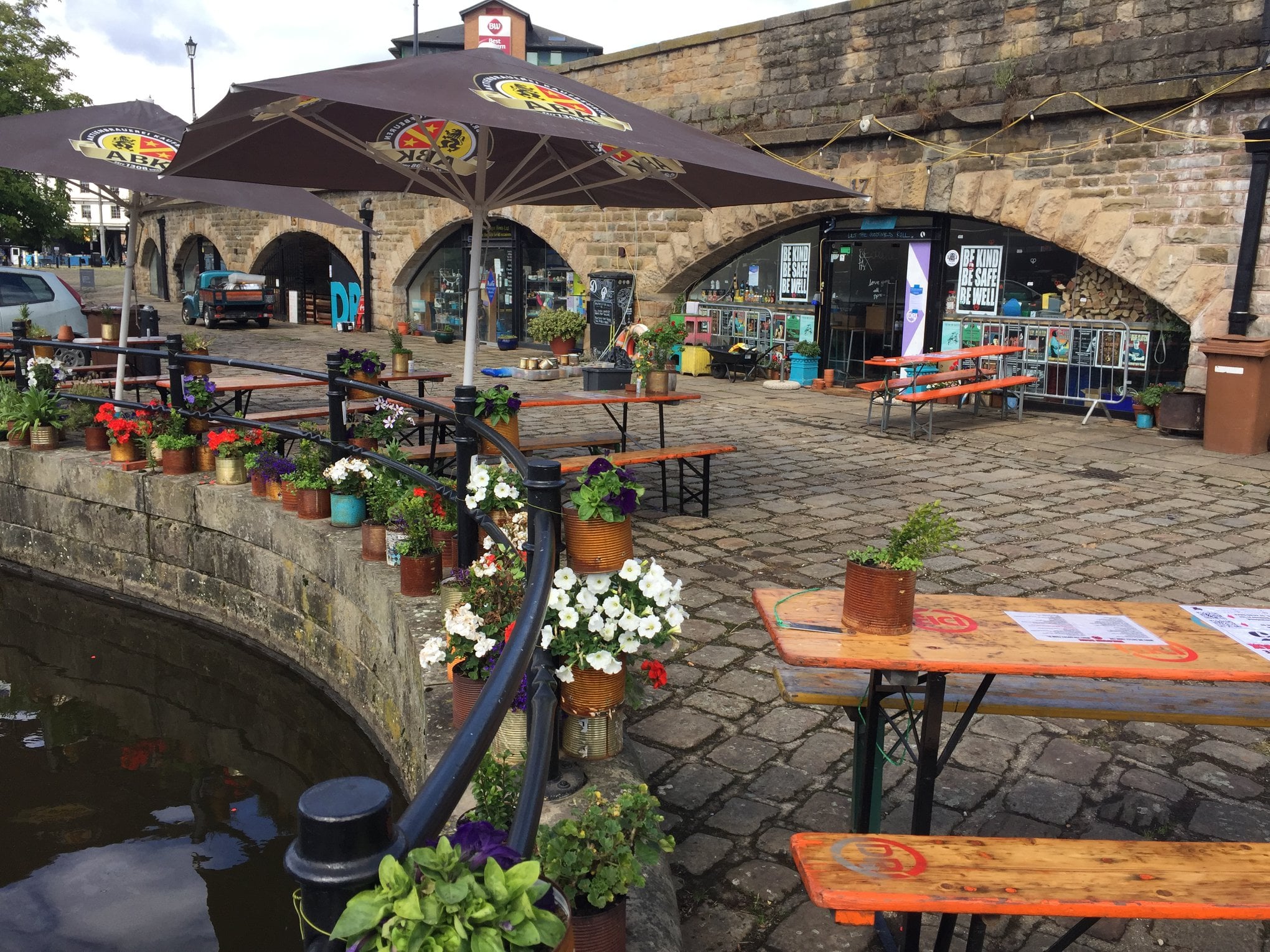 Outdoor seating area in front of a stone building with large archways. The space features wooden picnic-style tables with orange tops & green metal legs, some under large branded umbrellas. Colourful potted flowers line the edge of a canal in the foreground, adding vibrant reds, whites, and purples. Behind the seating area, the building has signage and posters visible through the arches, suggesting a café or bar setting. The ground is paved with cobblestones, and the overall scene has a relaxed atmosphere.