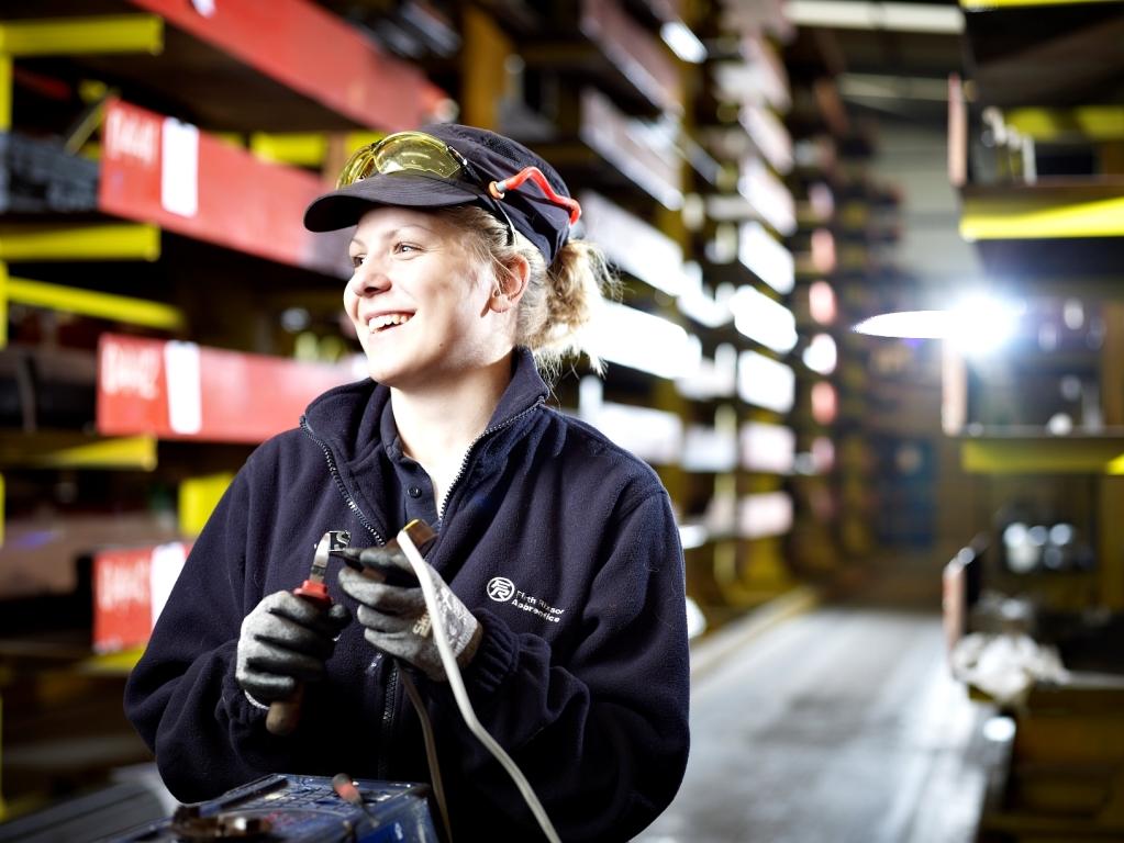 A person wearing protective work gloves and a dark industrial uniform stands in a warehouse aisle lined with metal shelving and stacked materials. The individual is holding a tool and a cable, with safety goggles resting on a cap. Bright lighting from the background highlights the organised rows of shelves and the industrial setting.