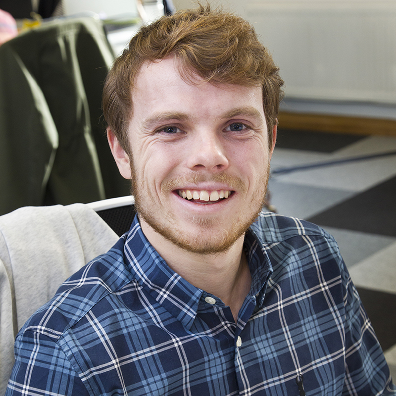 Person wearing a blue and green plaid button-up shirt, seated indoors on a chair with a light-coloured jacket draped over the back. The background shows a checked floor and part of a radiator along the wall.