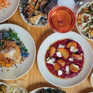 Top-down view of a wooden table set with several plates of colorful dishes, including a vibrant beetroot risotto topped with roasted golden beets and cheese, a plate of leafy greens with crispy garnish, and another with roasted meat and vegetables. Two glasses of wine—one white and one rosé—are also visible, along with small bowls of sides and garnishes.